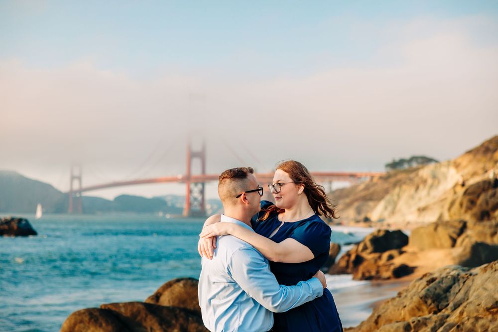 Couple embraces on rocky beach with Golden Gate Bridge in background during sunset in San Francisco.
