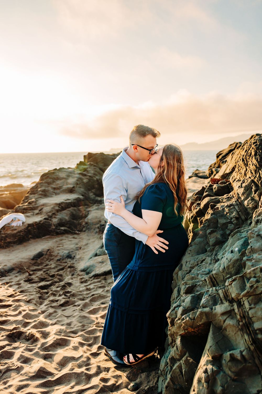Romantic sunset beach photoshoot with couple embracing by rocky coastal formations.