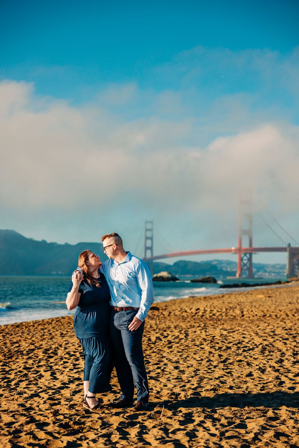 Couple enjoying a romantic moment on a sandy beach with the Golden Gate Bridge visible in the background.