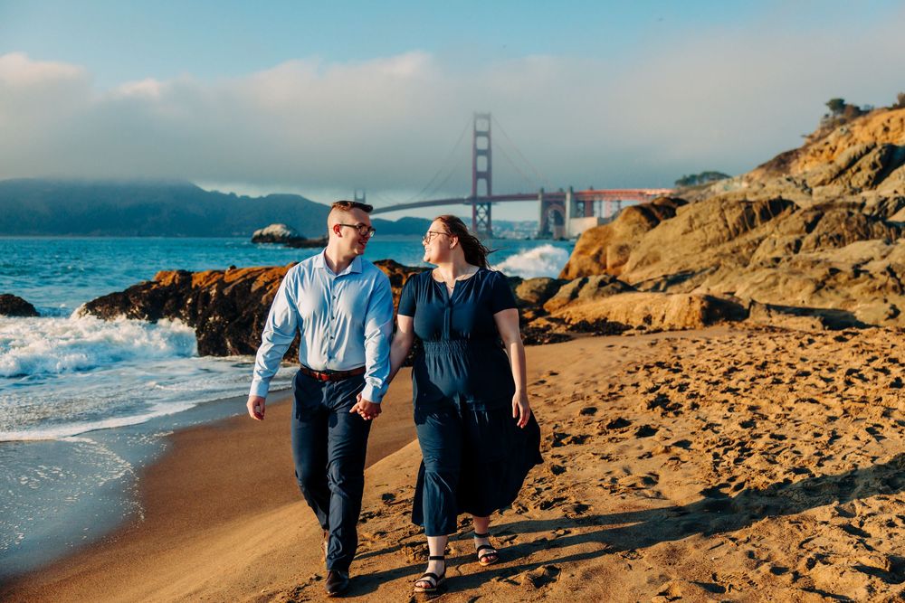 Couple walking along Baker Beach shoreline with dramatic views of Golden Gate Bridge and cliffs.
