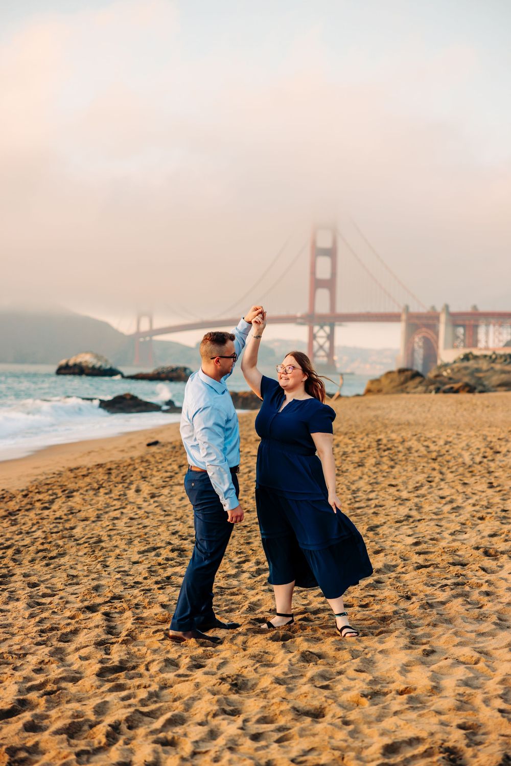 Couple poses playfully on sandy beach with Golden Gate Bridge visible in background at sunset.