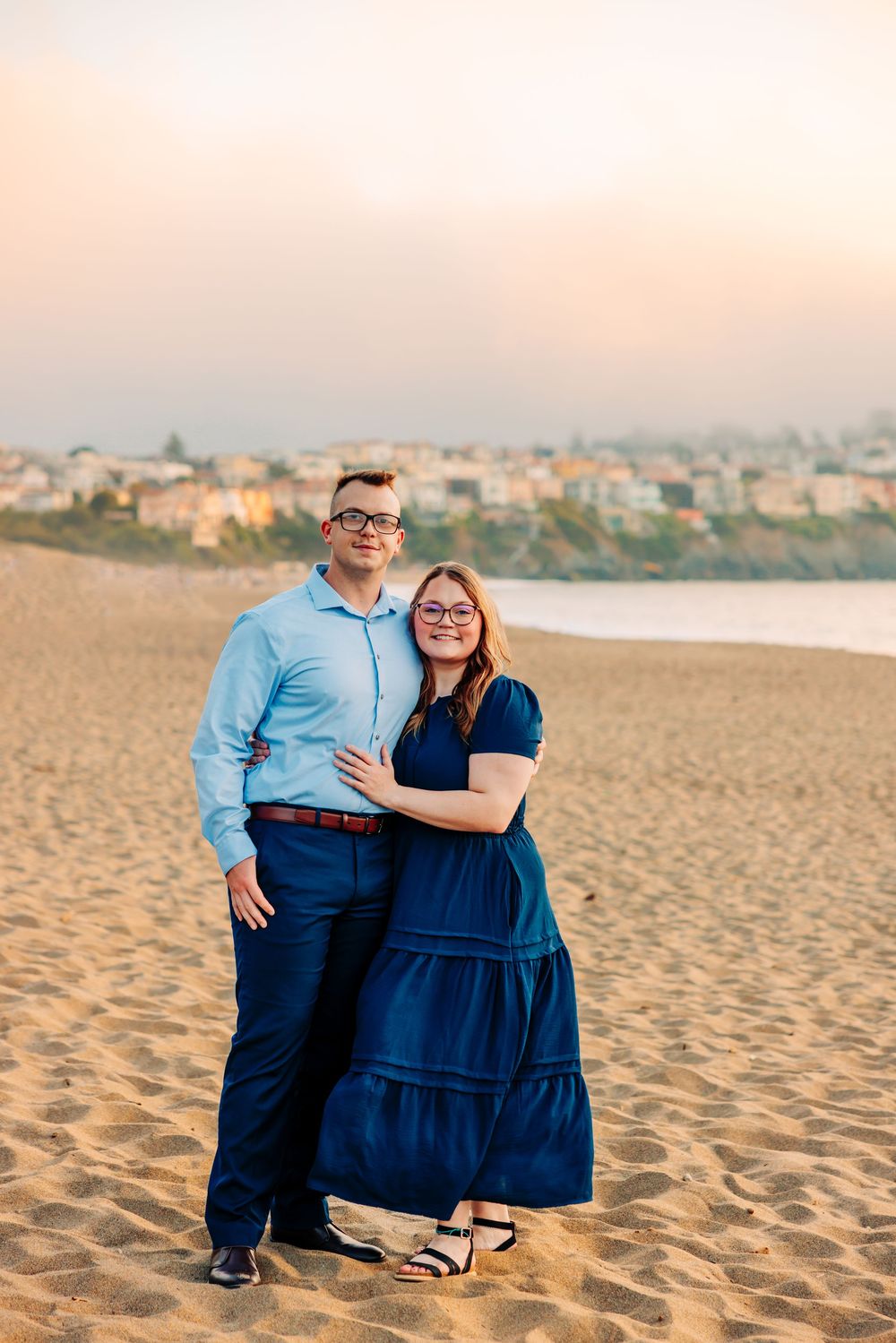 A couple poses together on a sandy beach at sunset with a coastal town visible in the background.