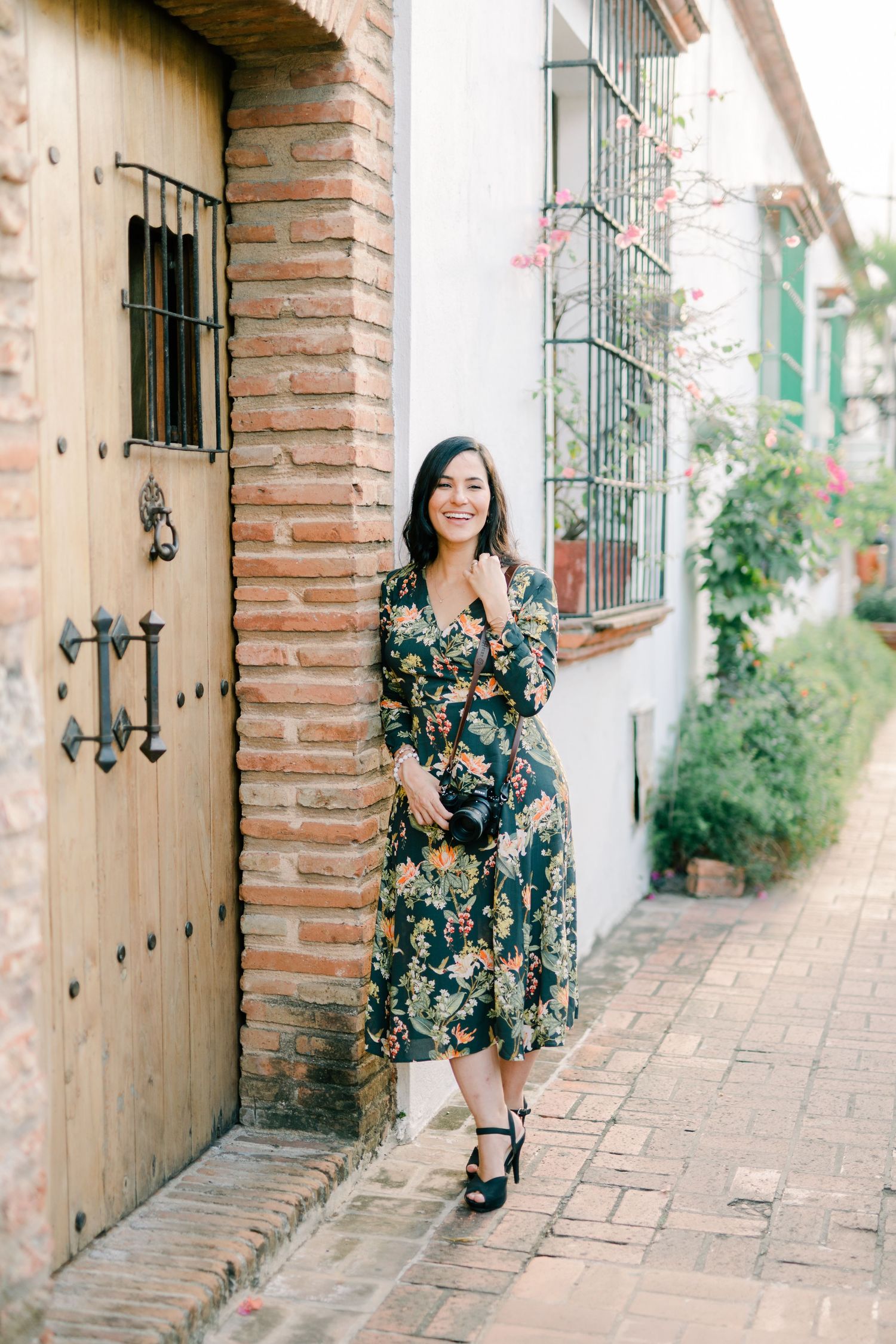 Professional photographer in white top and striped skirt smiles while holding vintage camera in outdoor garden setting.