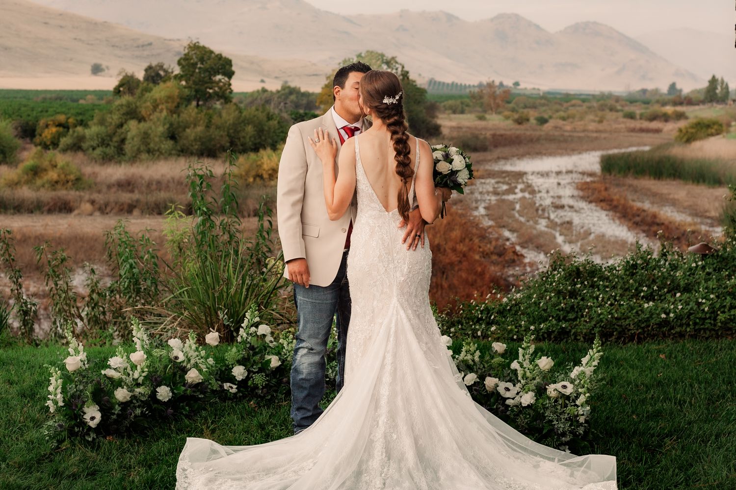 Bride in lace gown stands against mountain backdrop with floral arrangements.