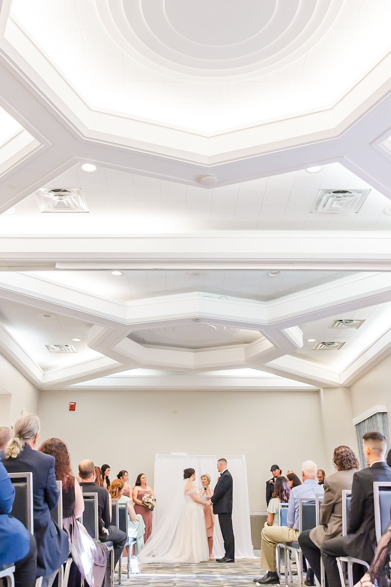 A panoramic view of a wedding reception venue with white octagonal ceiling details and guests mingling in formal attire.