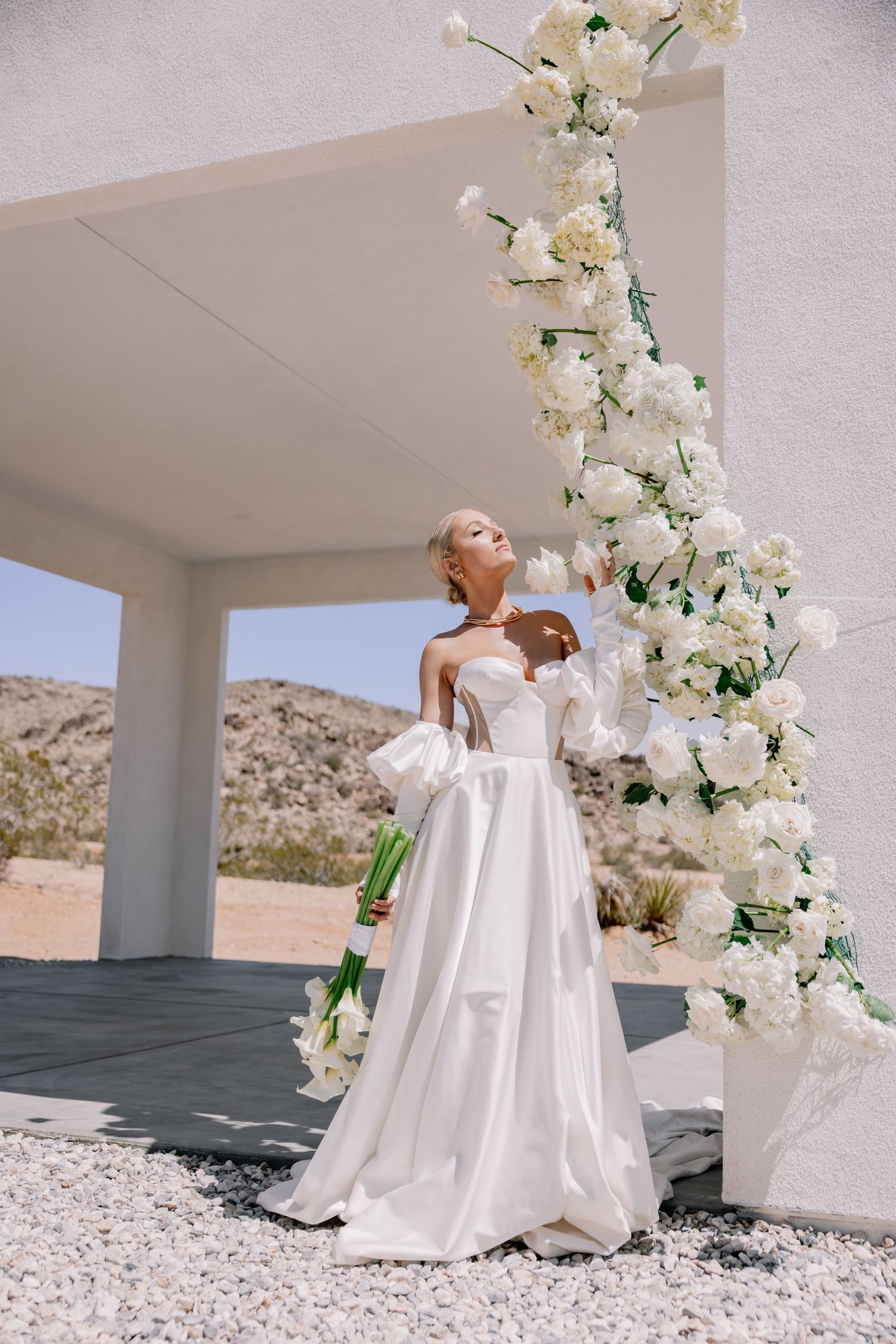 Elegant white wedding dress photographed in a desert setting with floral arch decorations.
