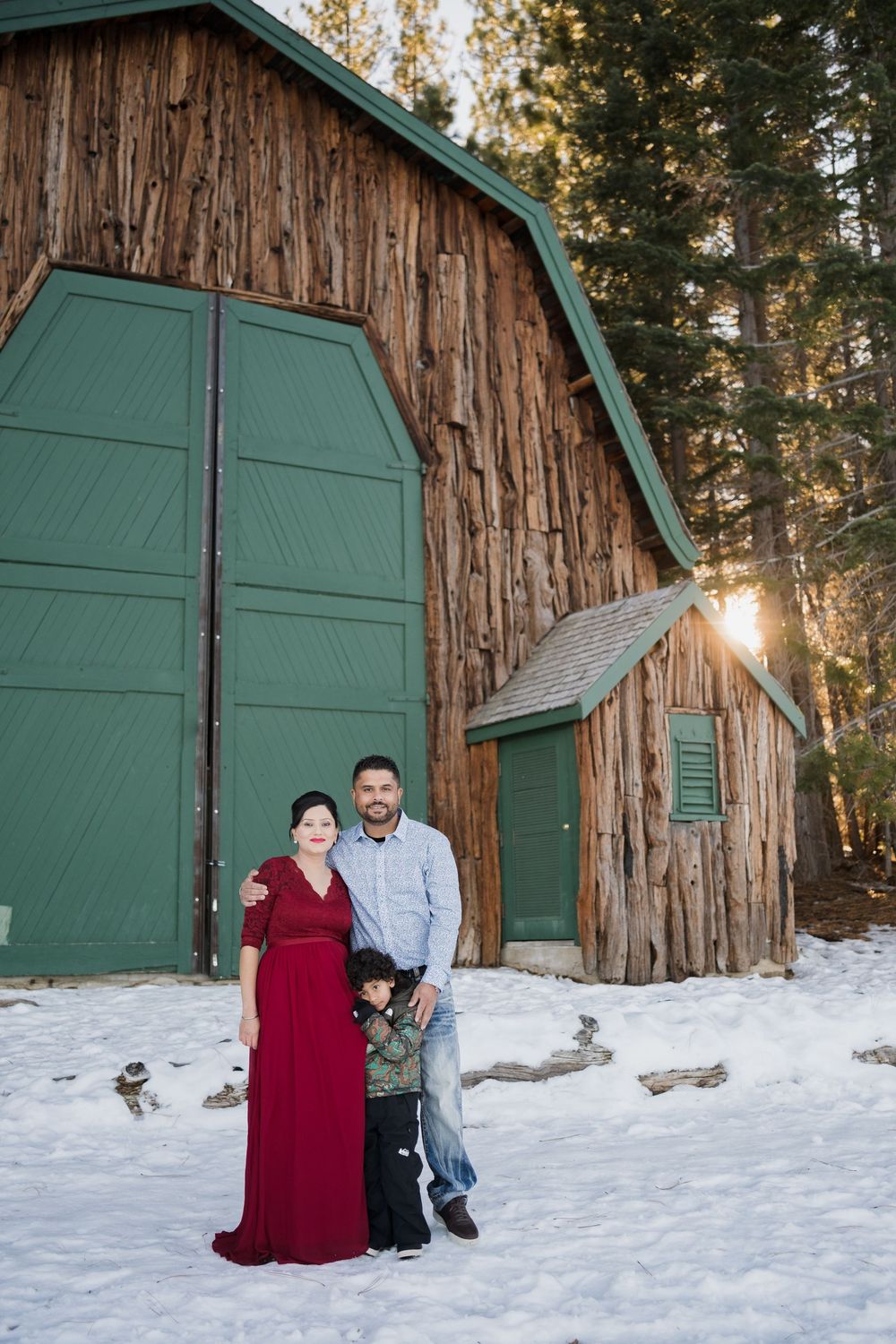 A couple poses together in front of a green wooden door against a large redwood tree trunk in snowy winter scenery.