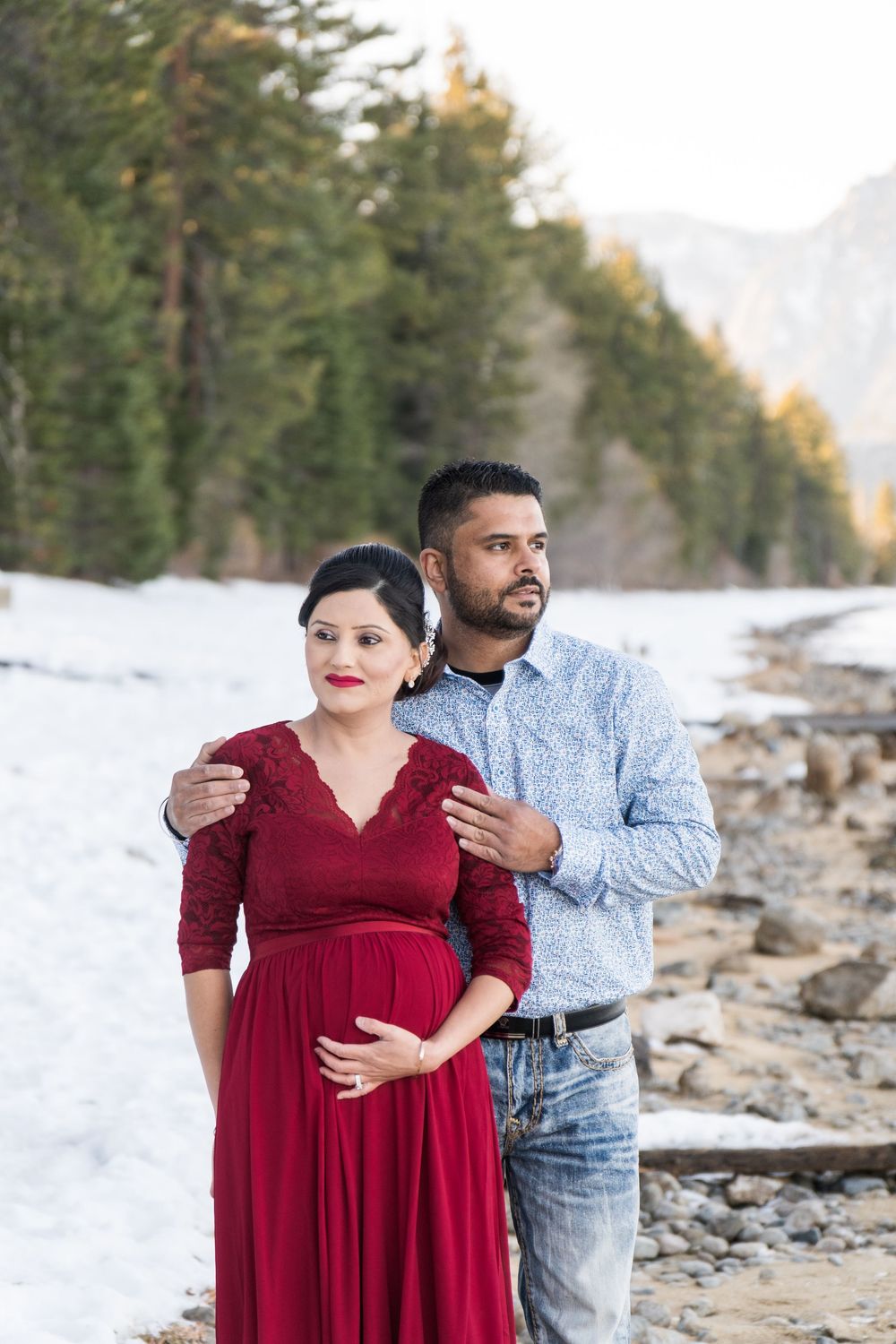 A couple poses together in a snowy mountain landscape, with evergreen trees and rocky terrain in the background.