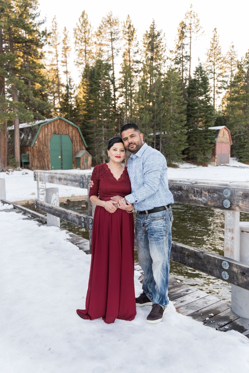 A couple poses together in winter by a snowy dock with pine trees and a rustic cabin in the background.