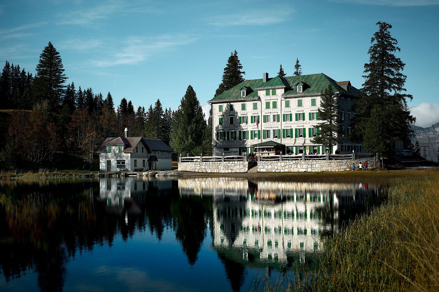 White lakeside lodge reflects perfectly in calm mountain waters surrounded by evergreen forest.