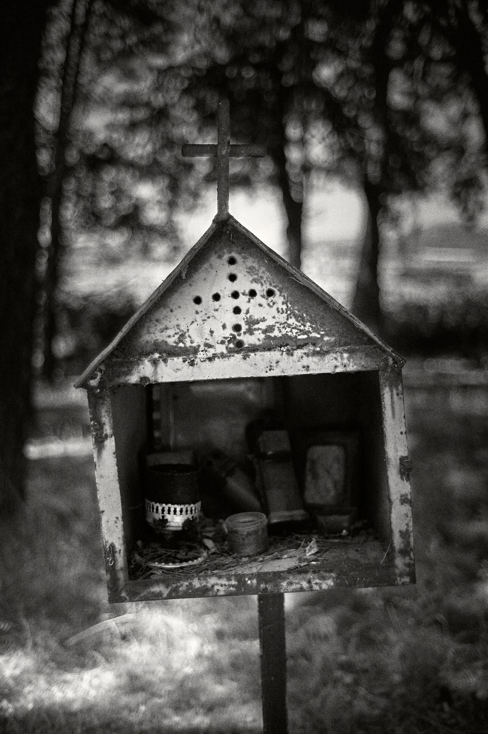 A rustic wooden birdhouse with a cross on top stands in black and white against a blurred forest background.
