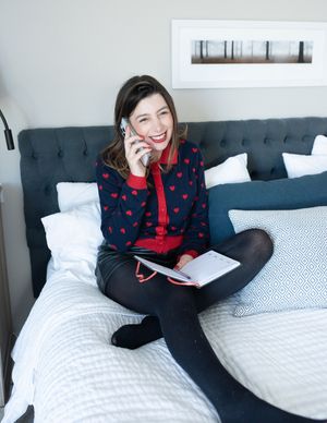 Smiling woman in heart-print cardigan sitting on bed talking on phone with notebook.