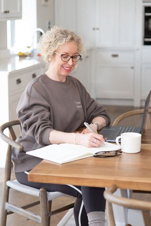Smiling middle-aged woman with curly blonde hair and glasses writing in notebook at wooden kitchen table with laptop.