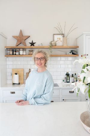 Smiling woman with glasses in light blue sweatshirt standing at white kitchen counter with open shelving and subway tile.