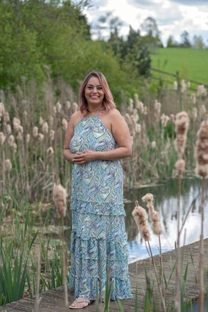 Woman in blue paisley halter maxi dress standing on wooden dock surrounded by cattails near a pond.