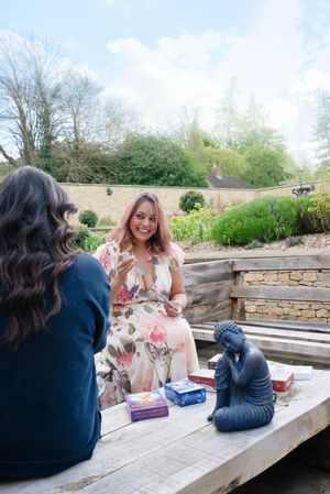 Two women sit outdoors at a wooden table with tarot cards and a Buddha statue in a garden setting.