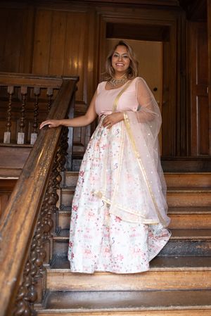 Woman in pink floral lehenga choli with dupatta smiling on wooden staircase in elegant interior.