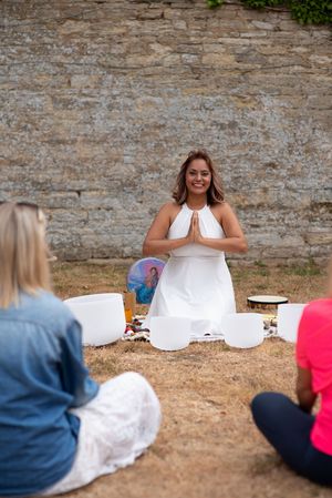 Woman in white dress meditates with prayer hands before crystal singing bowls outdoors, leading a sound healing session.