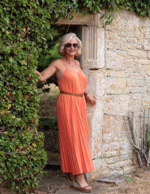 Older woman in orange pleated maxi dress with belt and sunglasses posing by stone wall with ivy.