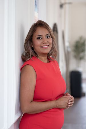 Smiling pregnant woman in red sleeveless dress leaning against white wall in bright hallway.
