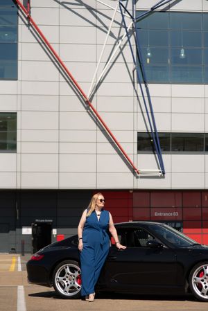 Woman in blue jumpsuit posing beside a black Porsche sports car outside a modern building with colorful geometric design.