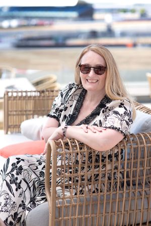 Smiling woman with blonde hair and sunglasses relaxing in a wicker chair outdoors, wearing a black and white floral dress.