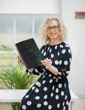 Woman in polka dot dress reads from black folder while sitting near indoor plants.