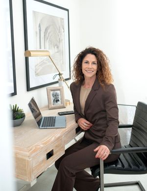 Professional in brown suit sitting in modern office with laptop and framed artwork.
