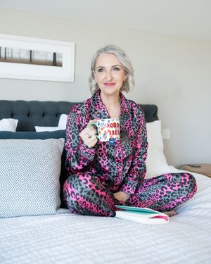 Woman in pink leopard print satin pajamas sitting cross-legged on bed holding colorful mug with book nearby.