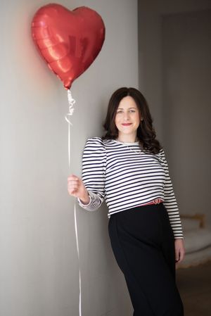 Person in striped shirt poses with red heart balloon against grey background.