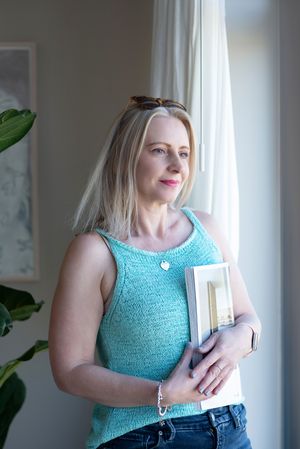 A person in a turquoise tank top holds books while standing by a bright window.