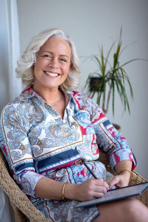 Woman in patterned blouse sits in wicker chair writing in notebook with houseplants in background.