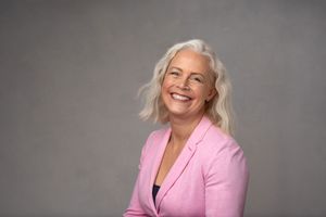Professional wearing a pink blazer smiles warmly against a grey studio backdrop.
