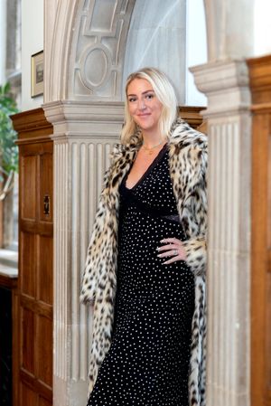 Woman in black polka dot dress and leopard print coat poses against ornate architectural backdrop in elegant setting.