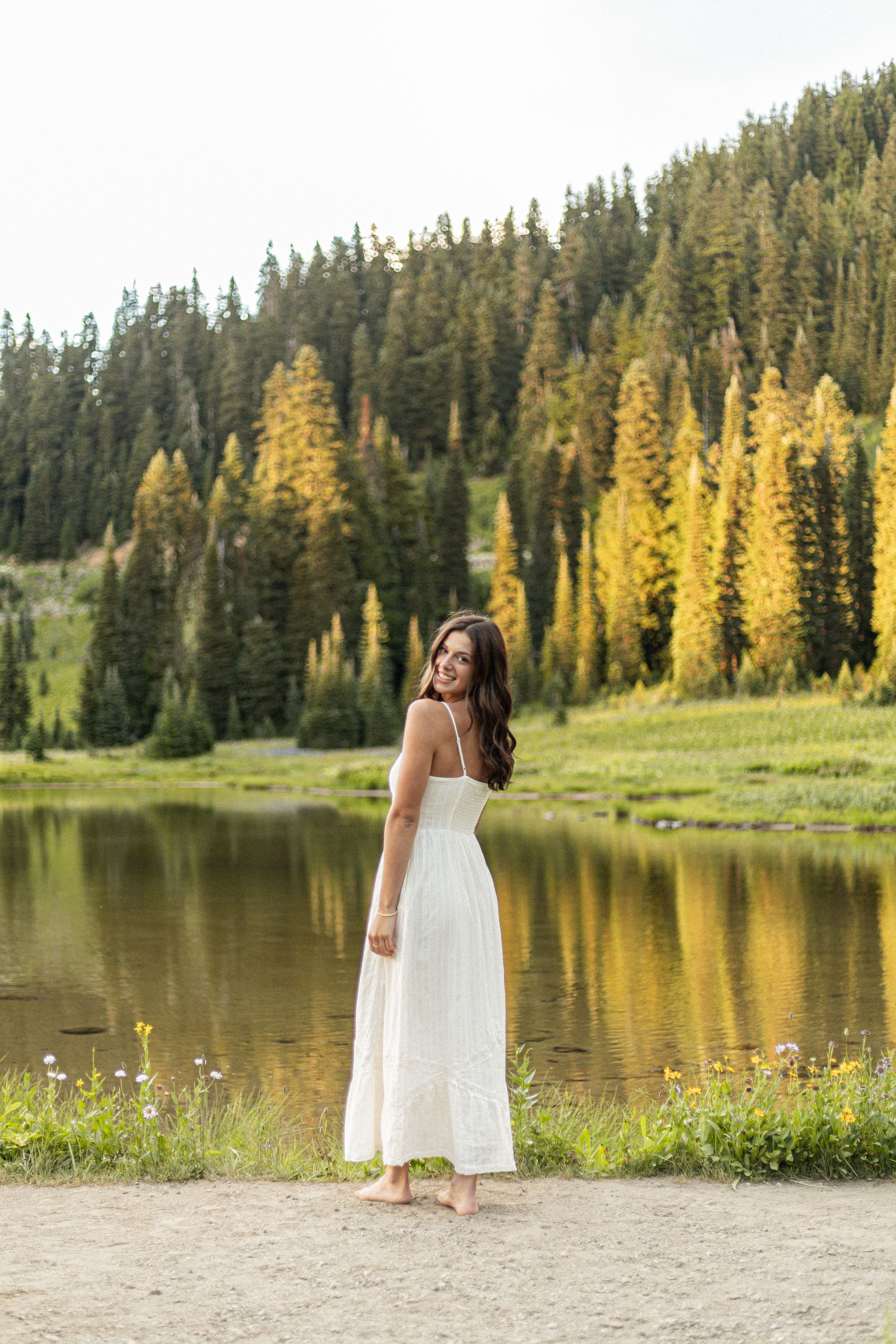 A figure in a flowing white dress stands barefoot by a serene mountain lake surrounded by evergreen and golden trees.
