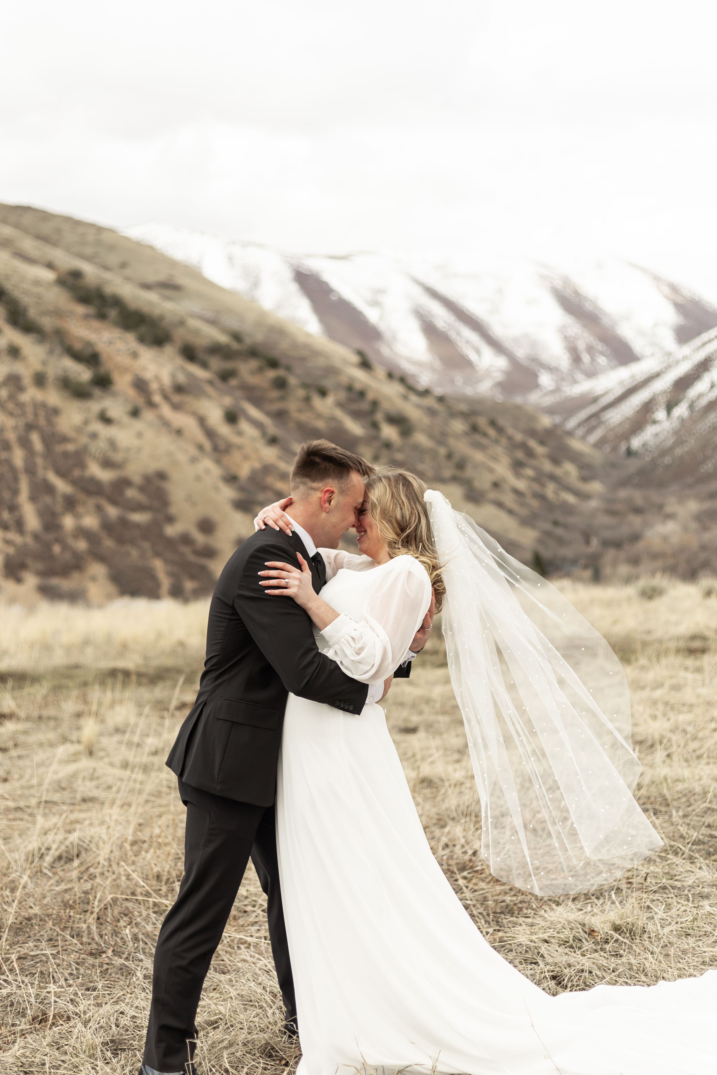 A romantic wedding couple embraces in a scenic mountain landscape with a flowing white veil in the breeze.