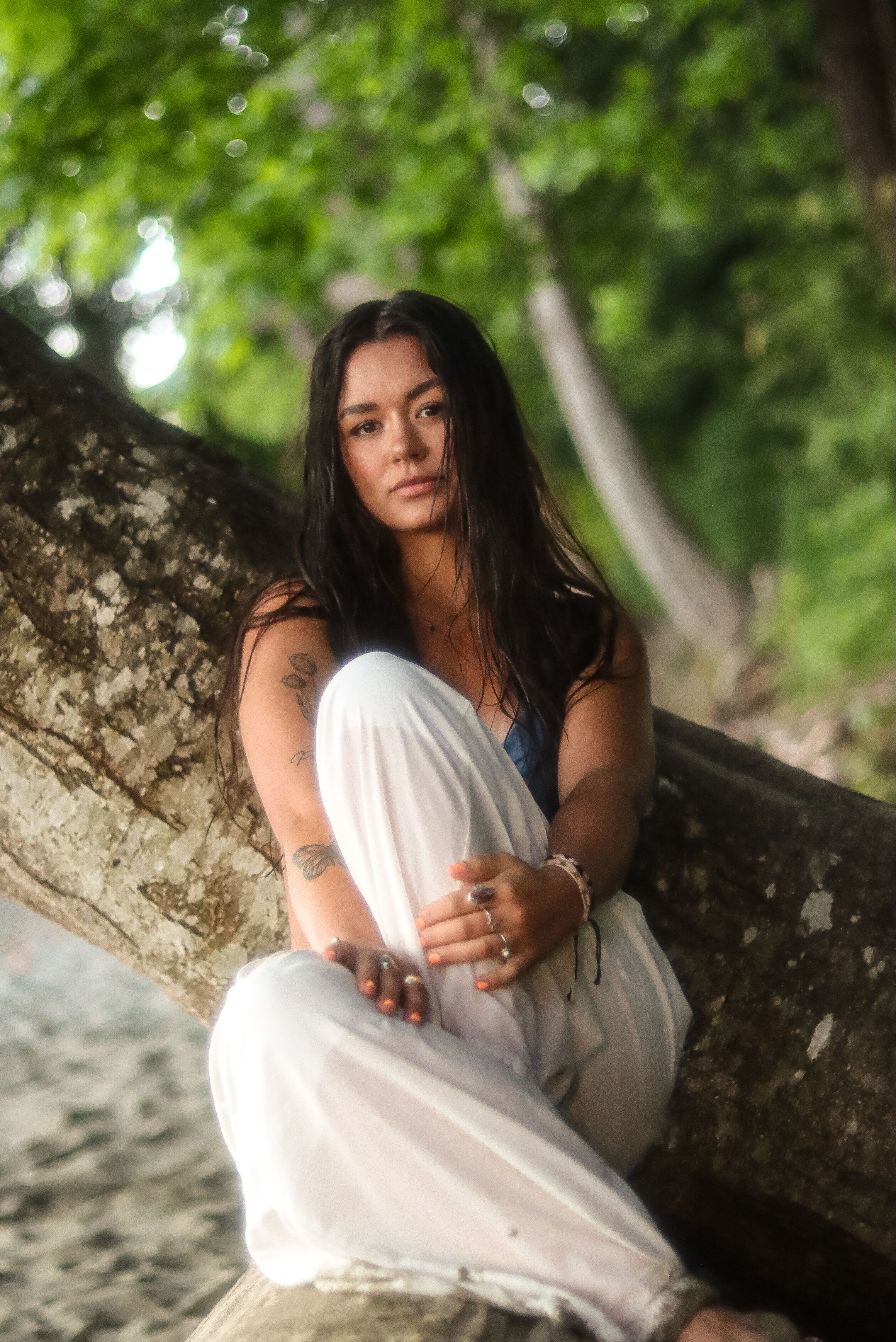 A person in a flowing white dress poses gracefully against a tree trunk on a beach with lush greenery in the background.