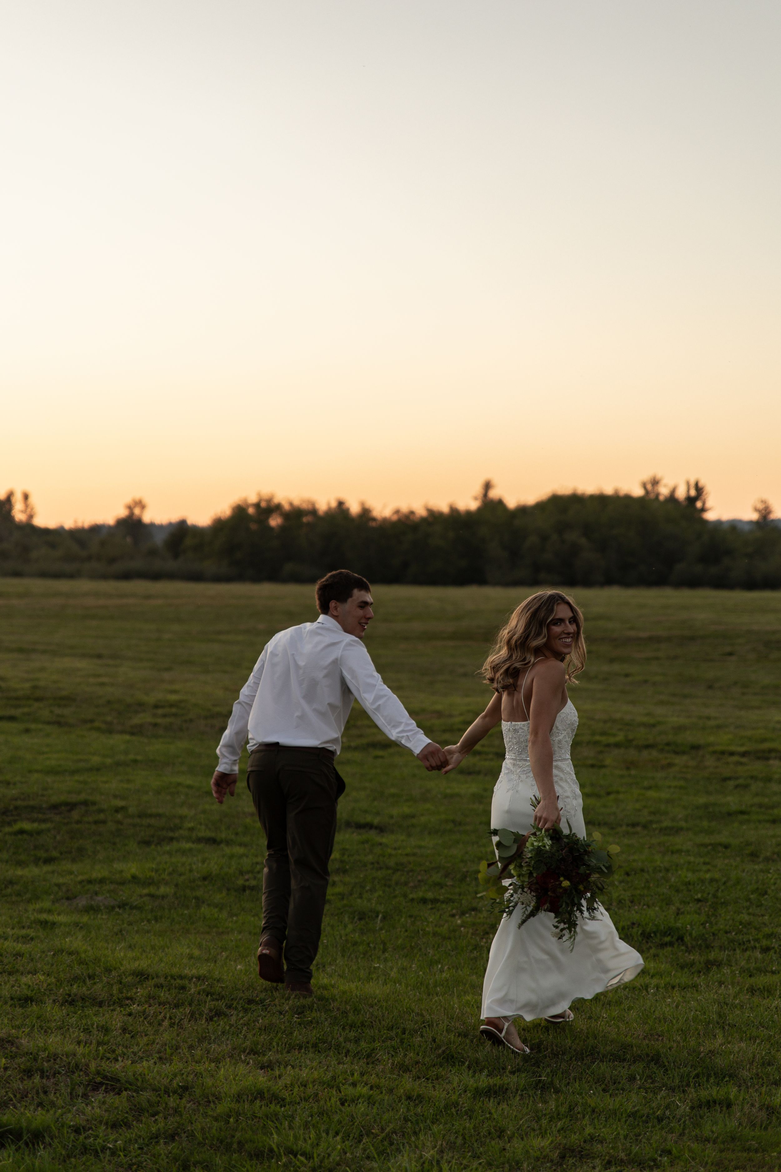 A couple runs playfully through a grassy field at sunset, with the bride holding her wedding bouquet.