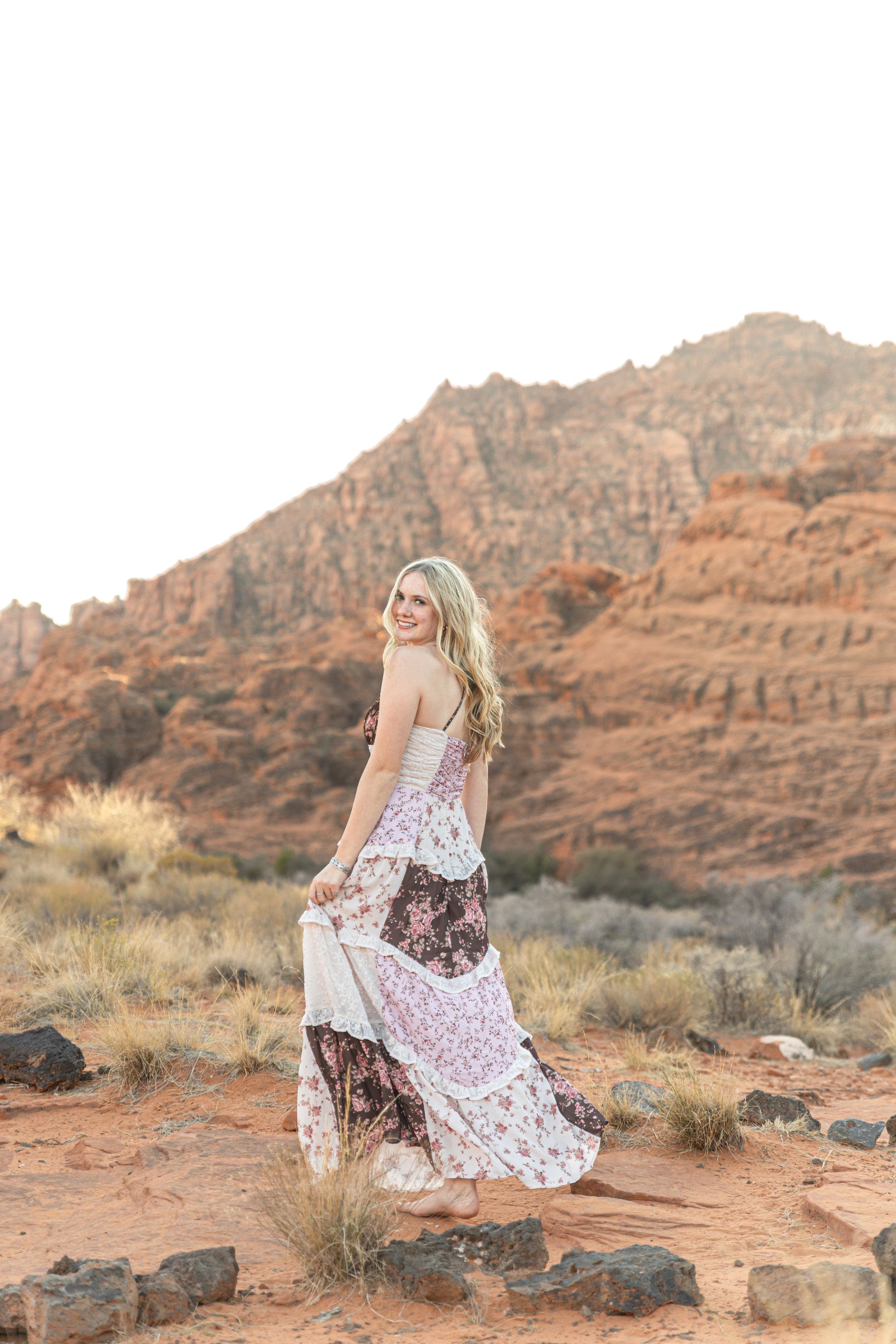 A flowing floral dress catches the wind against red rock formations in a desert landscape at sunset.