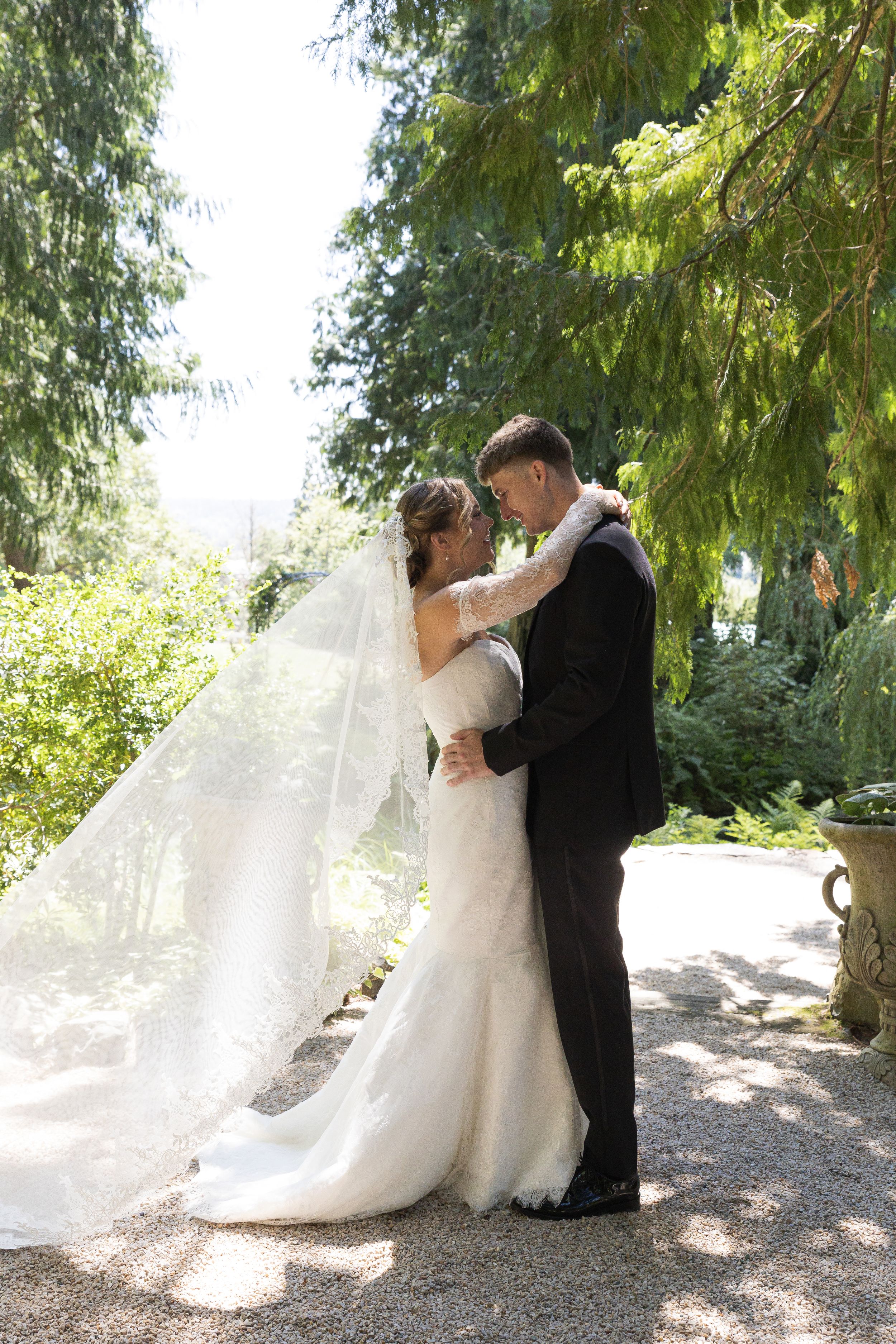 A romantic wedding couple embraces under evergreen trees in a garden setting as the bride's veil flows in the breeze.