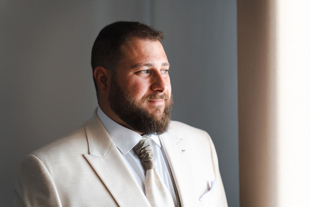 Bearded groom in cream suit gazing thoughtfully out a window in soft natural light.
