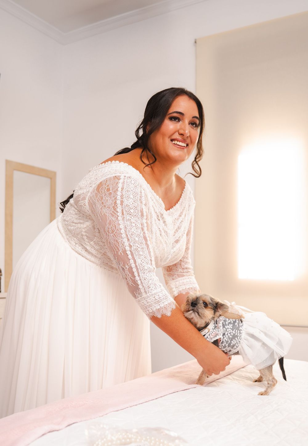 Smiling bride in lace wedding dress petting a small dog dressed in silver outfit on a bed.