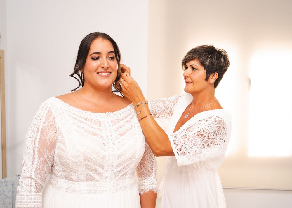 Bride in lace off-shoulder gown smiling as a woman adjusts her earring in a bright room.