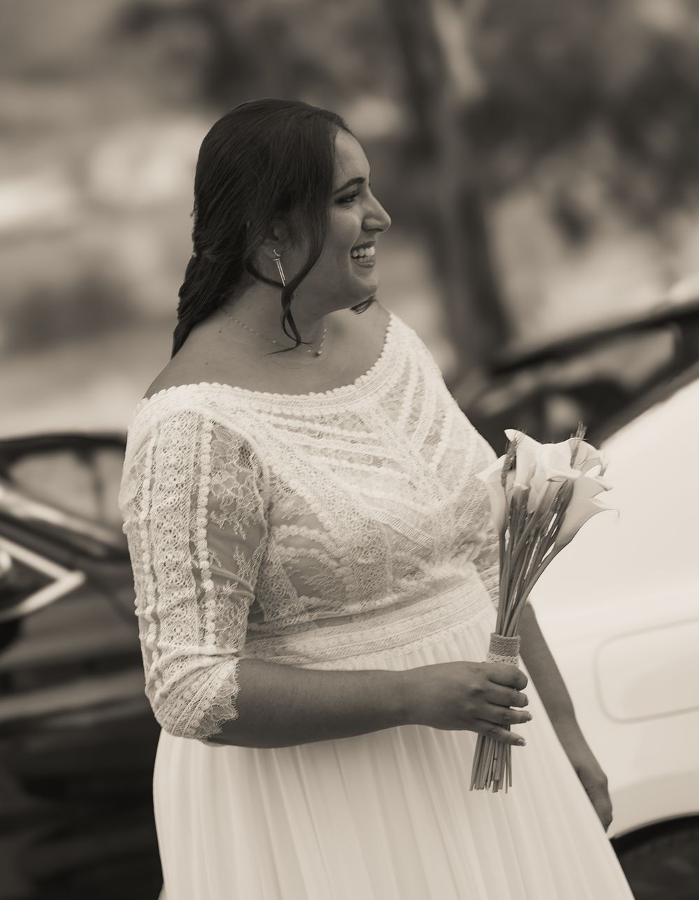 Black and white portrait of a smiling bride in lace dress holding white calla lilies outdoors.