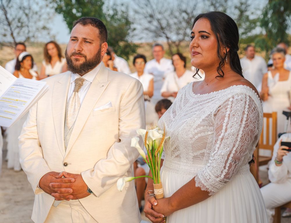 Bride and groom standing together at outdoor beach wedding ceremony with white-dressed guests behind them.