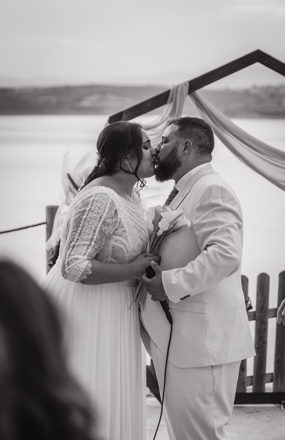 Black and white photo of bride and groom sharing first kiss at outdoor waterfront wedding ceremony.