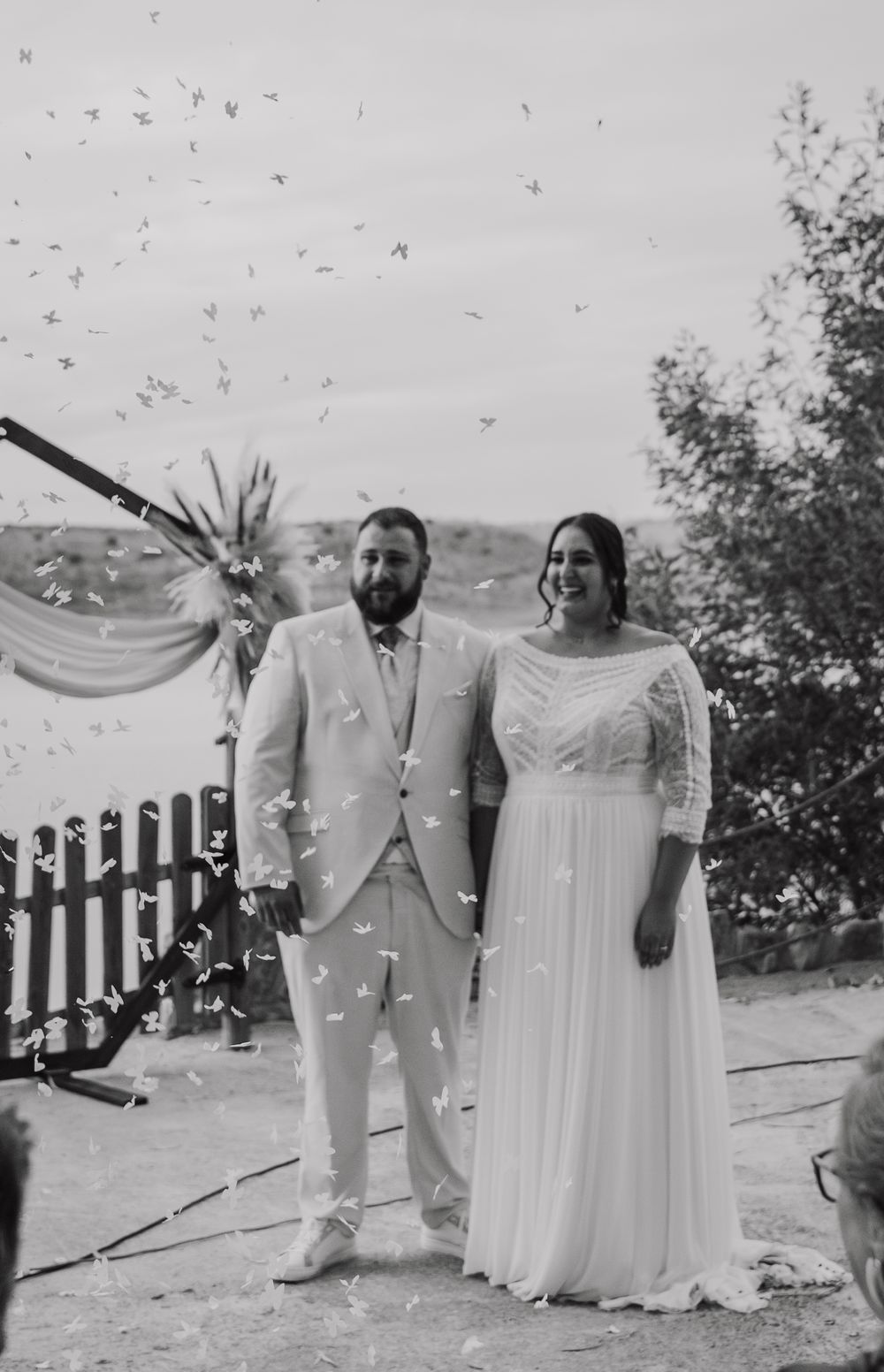 Black and white photo of a smiling bride and groom standing together as confetti falls during their outdoor wedding ceremony.