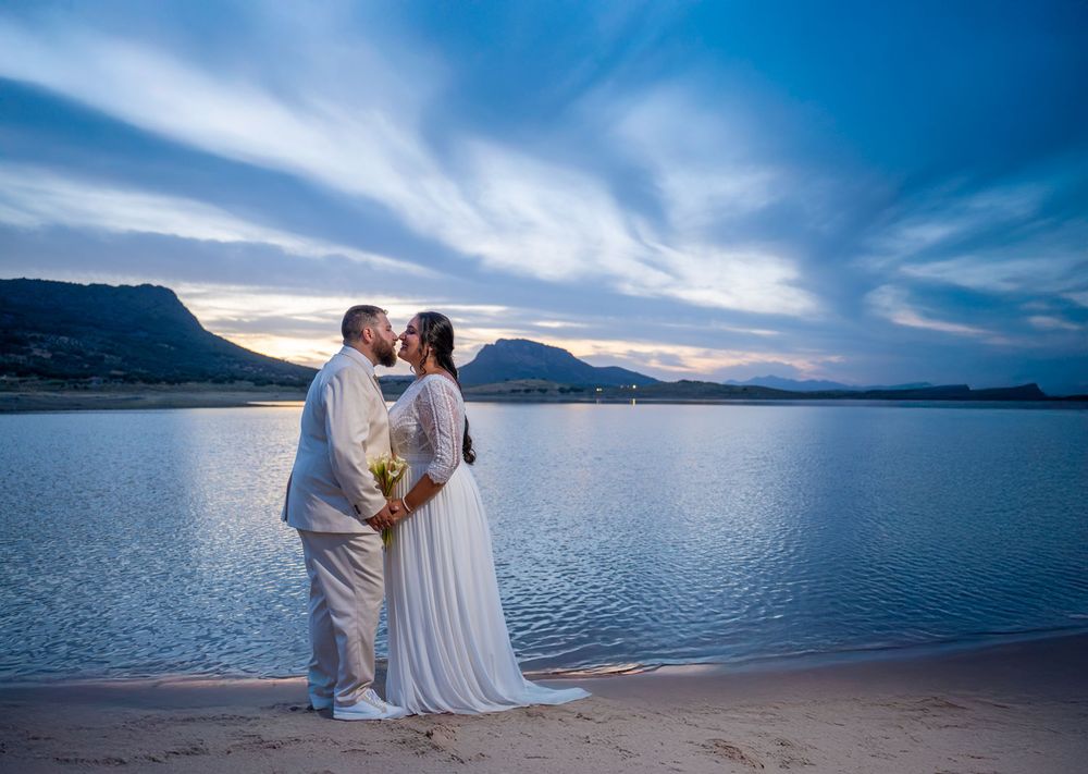 Bride and groom sharing a romantic kiss on a sandy beach at sunset with a stunning lake and mountain backdrop.