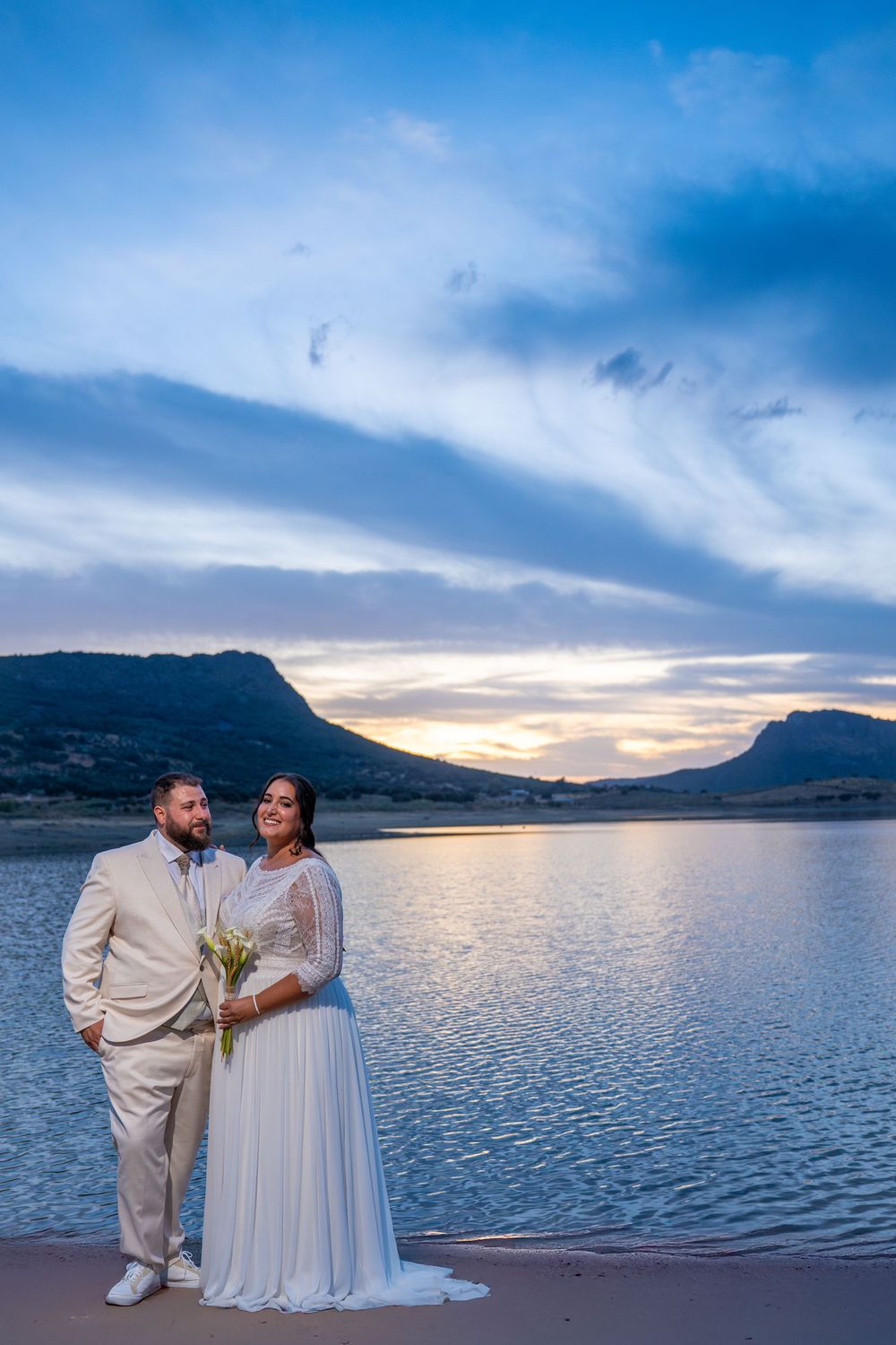 Bride holding a bouquet and groom standing together on a beach at sunset with a serene lake and mountain view.