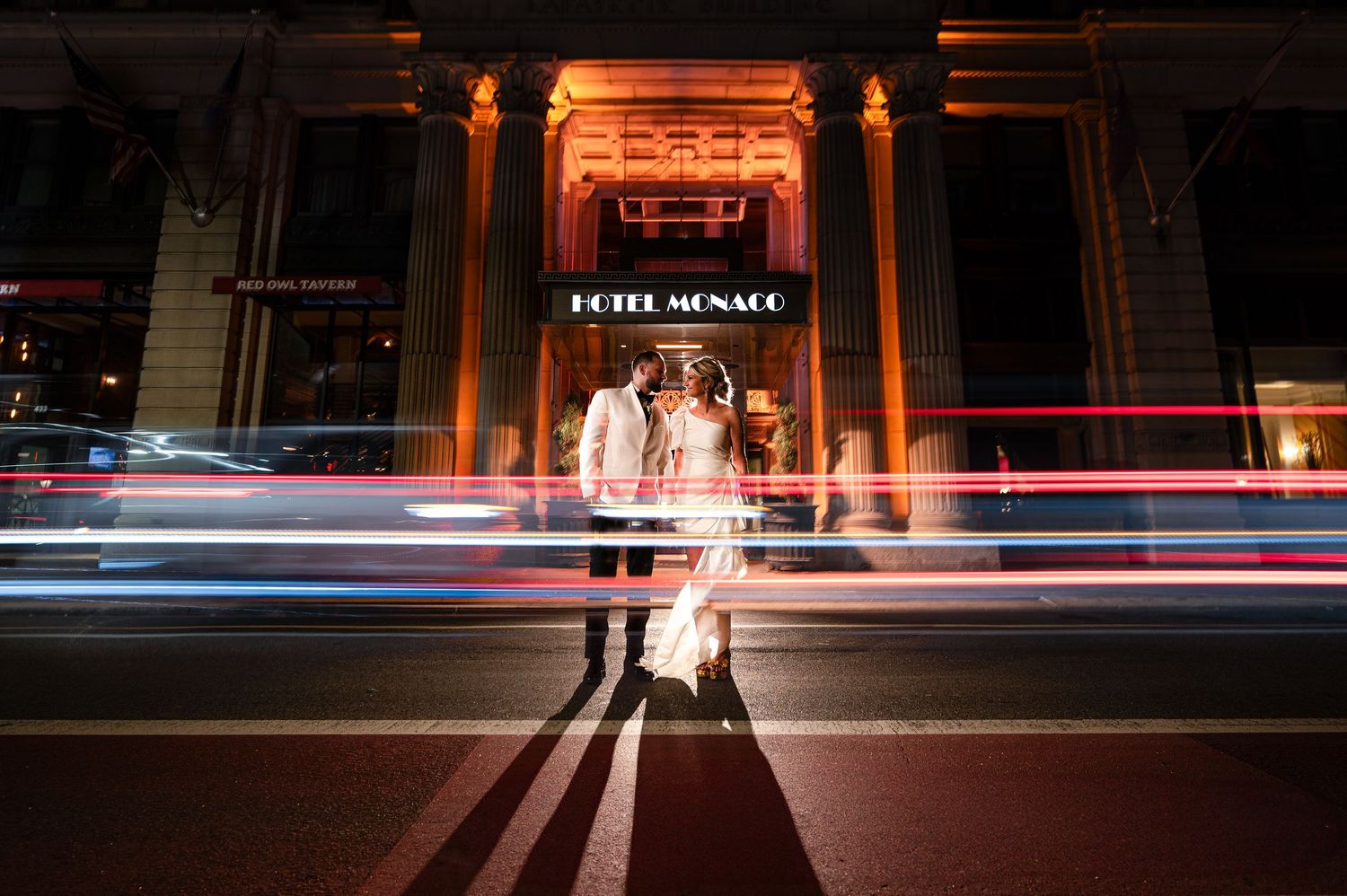 Long exposure nighttime photo showing red light trails and illuminated storefronts along a city street.